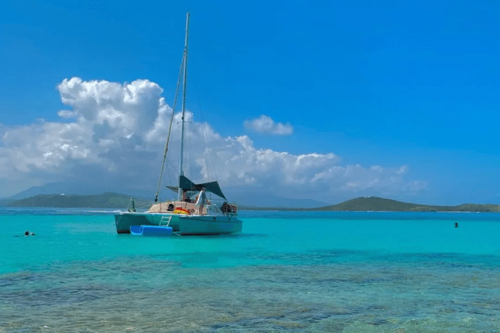 Sailboat anchored in turquoise sea with distant hills under a bright blue sky with clouds.
