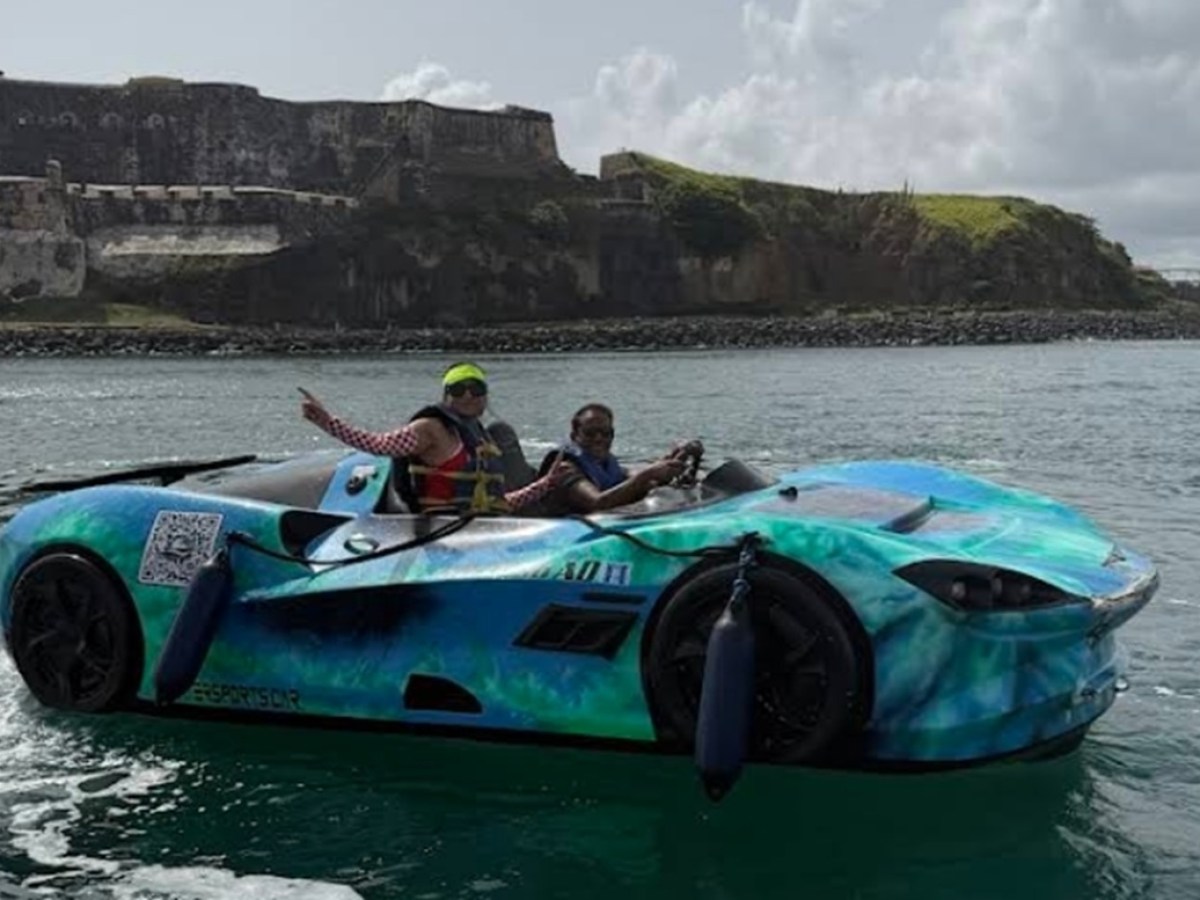 Two people in a boat shaped like a sports car on water, with a fort in the background.