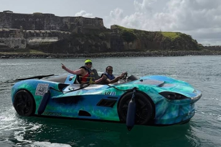 Two people in a boat shaped like a sports car on water, with a fort in the background.