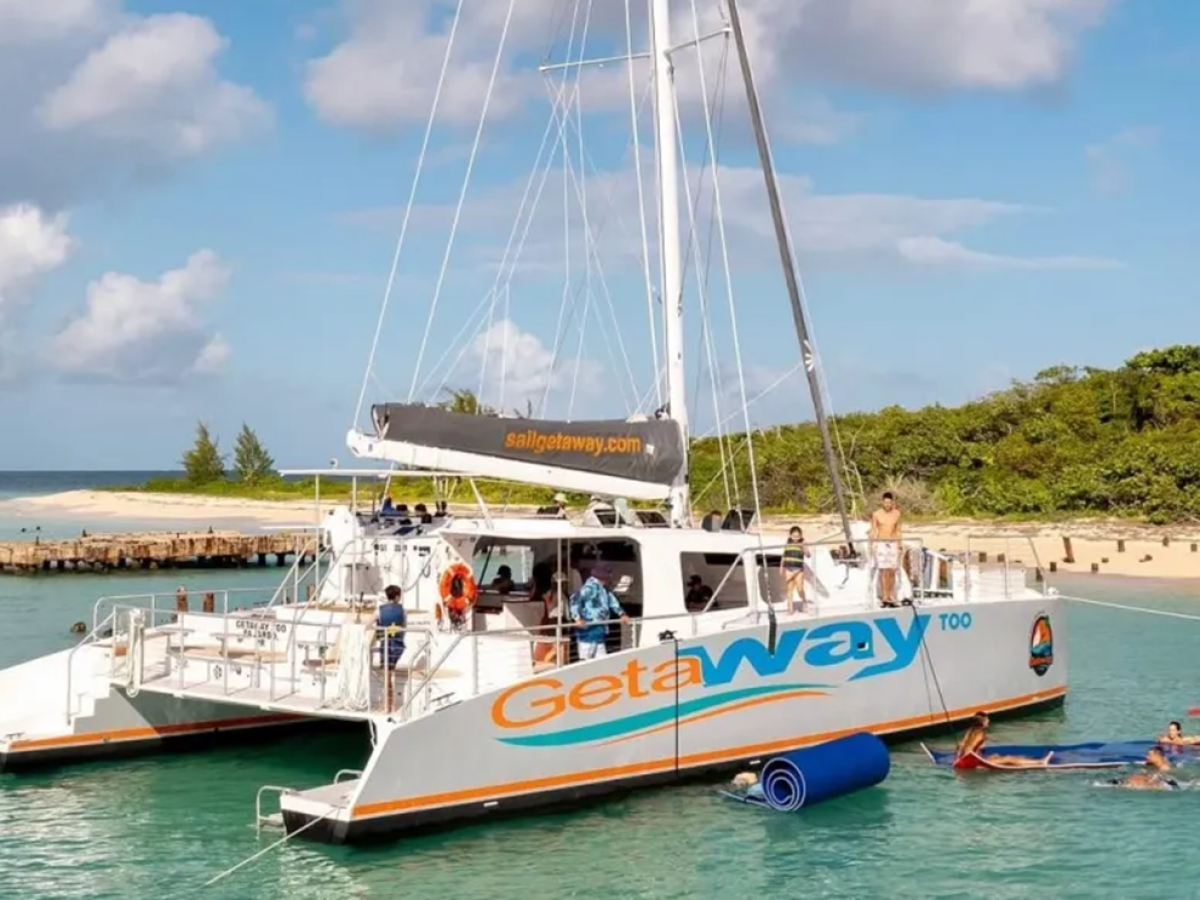 Large catamaran with people relaxing near a tropical island coast under a blue sky.