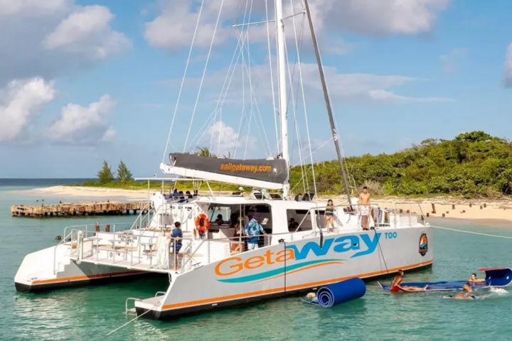 Large catamaran with people relaxing near a tropical island coast under a blue sky.