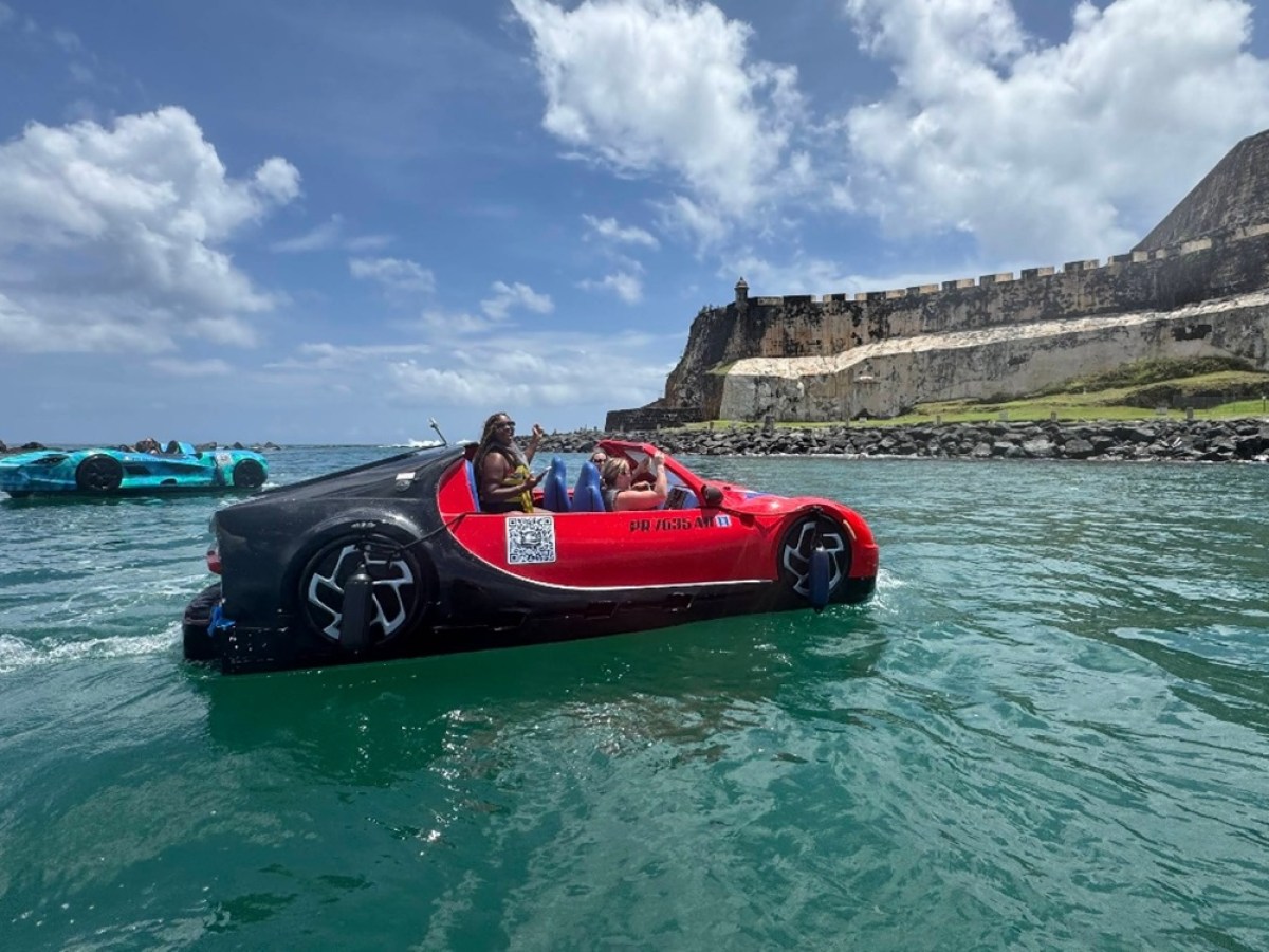 Floating car shaped boats on water near a historic fort under a partly cloudy sky.