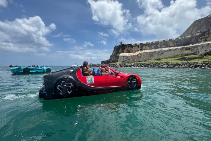 Floating car shaped boats on water near a historic fort under a partly cloudy sky.