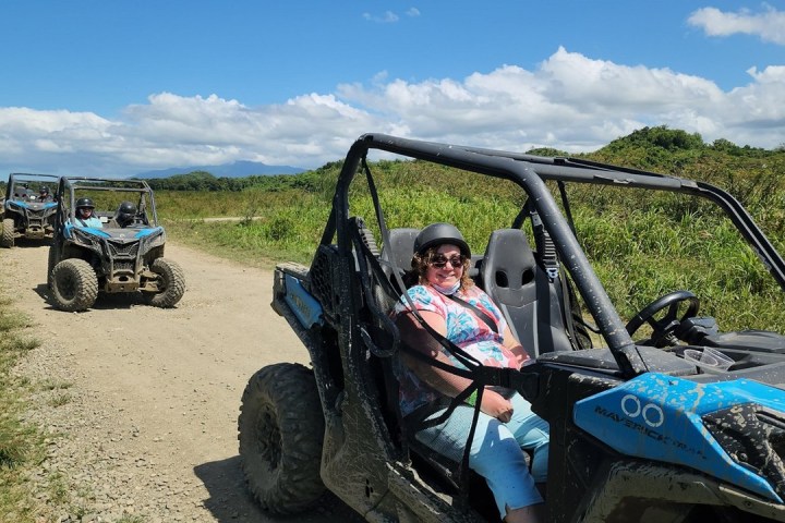 People driving off-road vehicles on a dirt path in a grassy landscape under a blue sky.