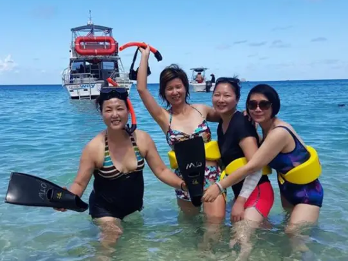 Four women in swimsuits with snorkeling gear in the ocean, a boat in the background under a clear sky.