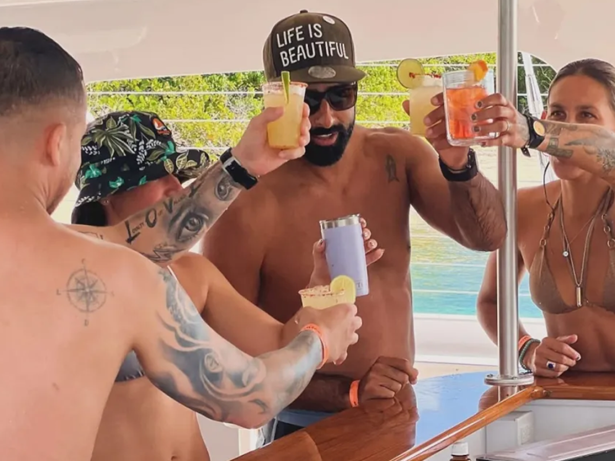 Group of people on a boat toasting with drinks, wearing swimwear and summer hats.