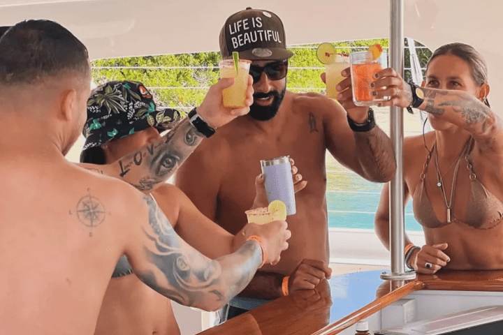 Group of people on a boat toasting with drinks, wearing swimwear and summer hats.