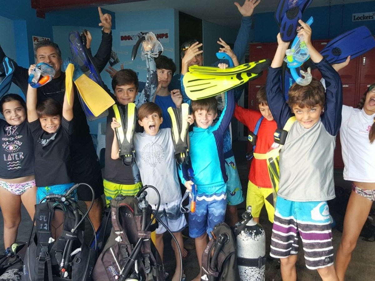 Group of excited kids and adult holding scuba gear, wearing colorful swimwear, smiling and cheering.