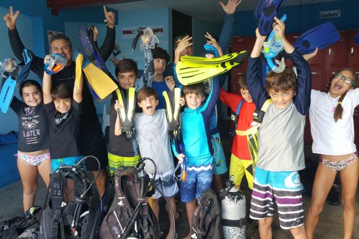 Group of excited kids and adult holding scuba gear, wearing colorful swimwear, smiling and cheering.