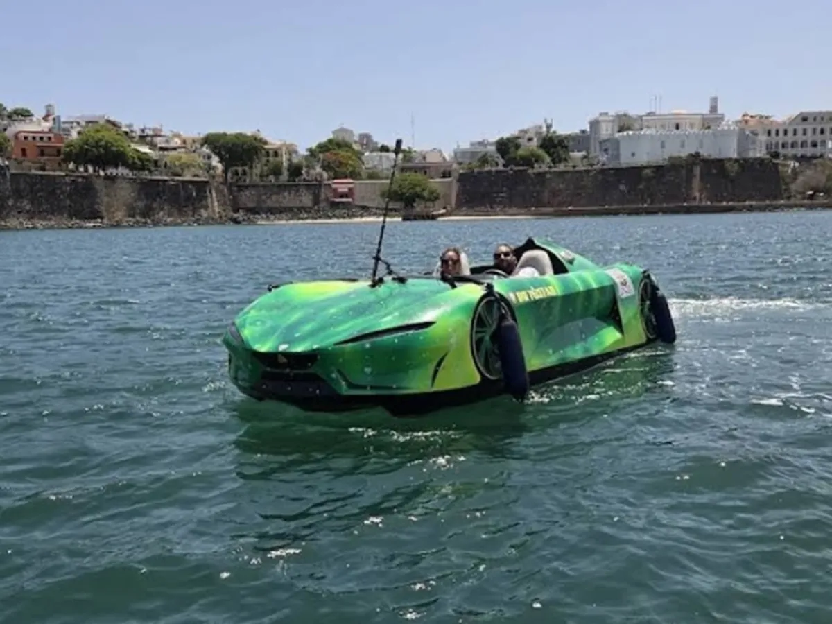 Green amphibious car with two passengers driving on water near a city shoreline.