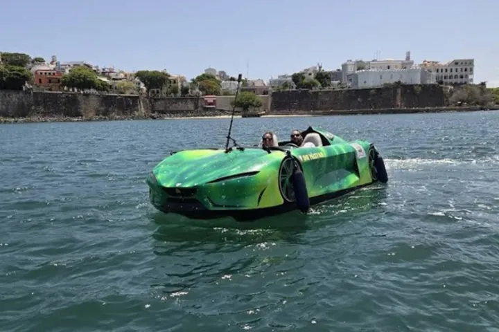 Green amphibious car with two passengers driving on water near a city shoreline.