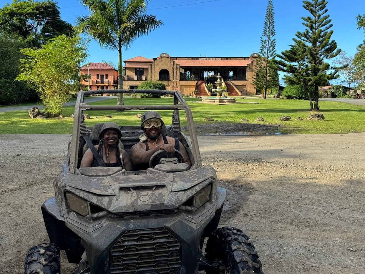 Two people smiling in a muddy off-road vehicle with a large house in the background.