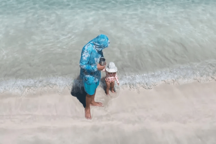 An adult and child in hats stand on a sandy beach by clear water.