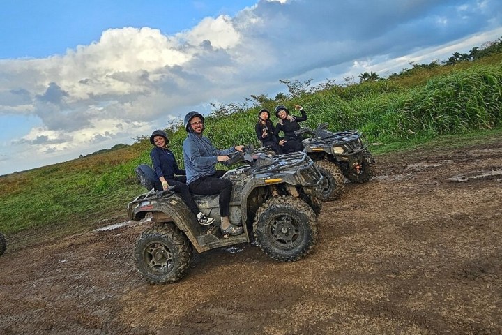 Three people on muddy ATVs posed on a dirt path with grass and cloudy sky in the background.