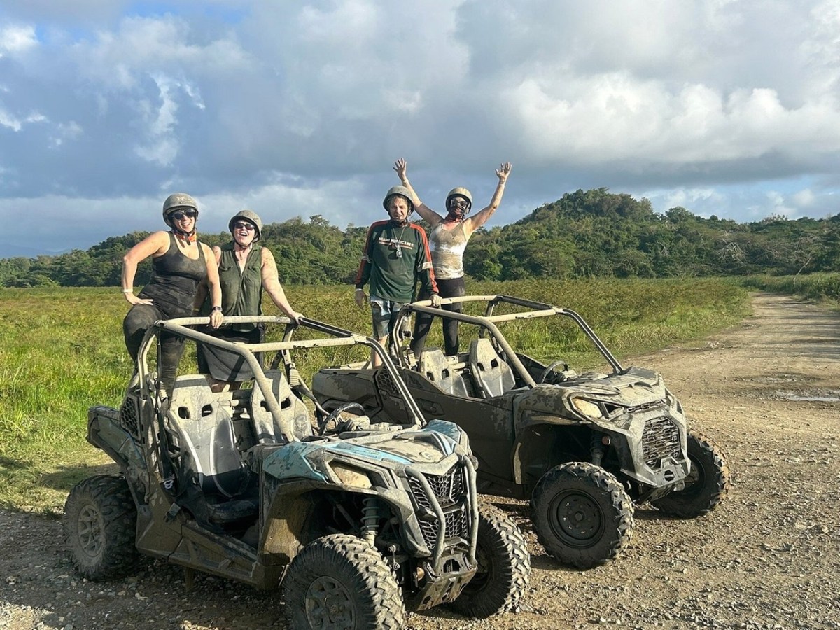 Four people in helmets pose with muddy ATVs in a grassy, open area with cloudy sky.
