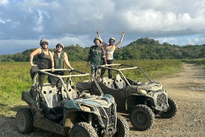 Four people in helmets pose with muddy ATVs in a grassy, open area with cloudy sky.