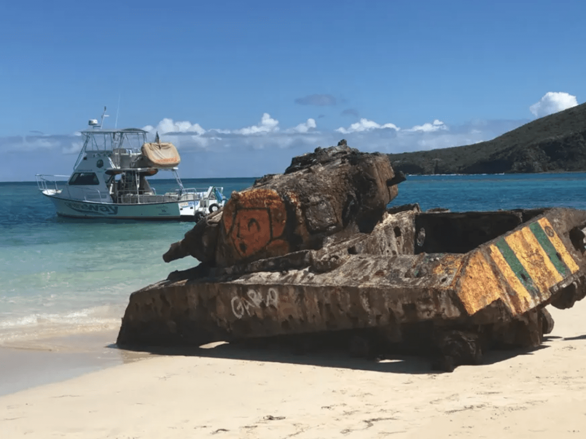 Rusty tank on a sandy beach with a boat and clear blue water in the background.