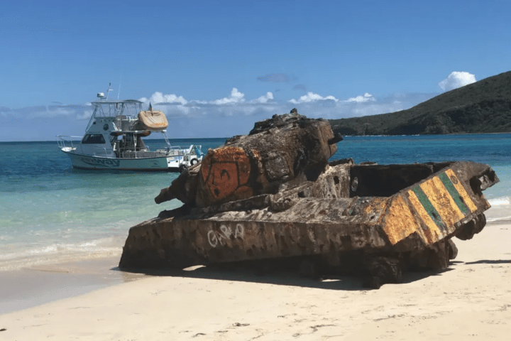 Rusty tank on a sandy beach with a boat and clear blue water in the background.
