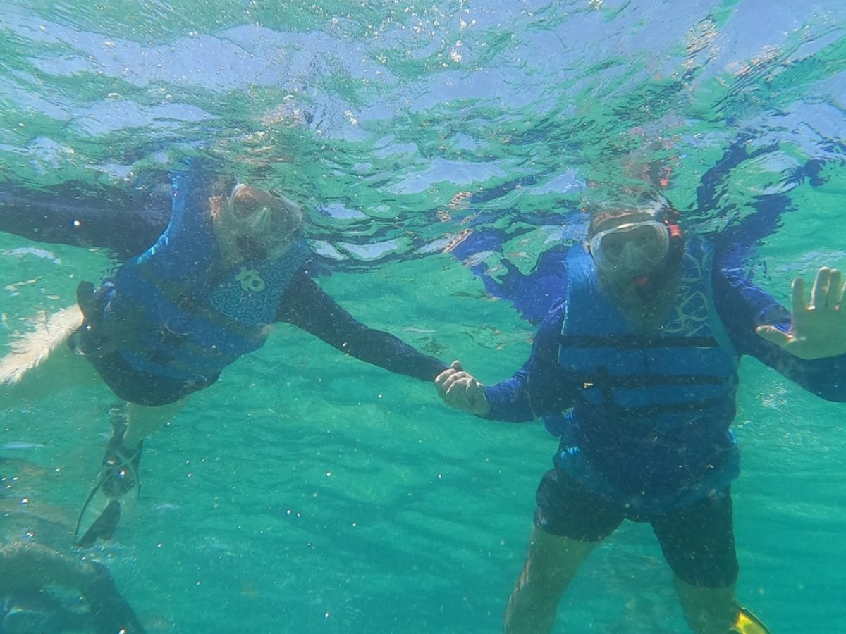 Two snorkelers in blue life vests underwater, holding hands, one waving.