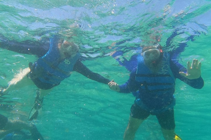 Two snorkelers in blue life vests underwater, holding hands, one waving.