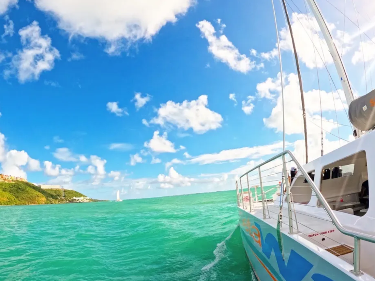 Sailboat on turquoise ocean near green hillside under partly cloudy blue sky.