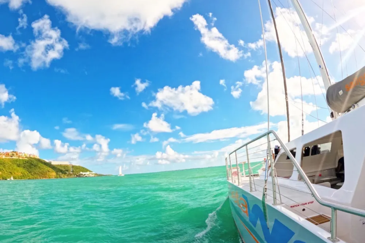 Sailboat on turquoise ocean near green hillside under partly cloudy blue sky.