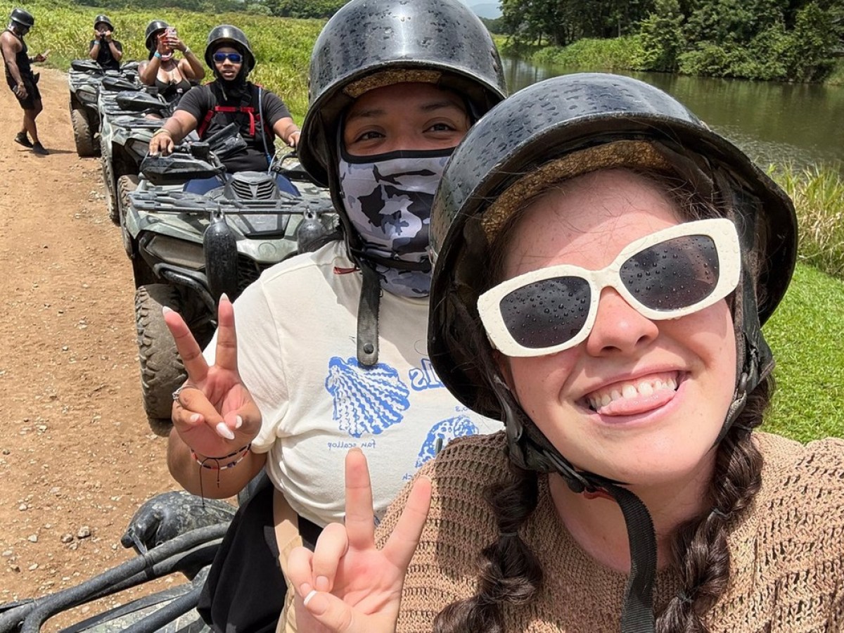 People wearing helmets on ATVs near a river, smiling and making peace signs.