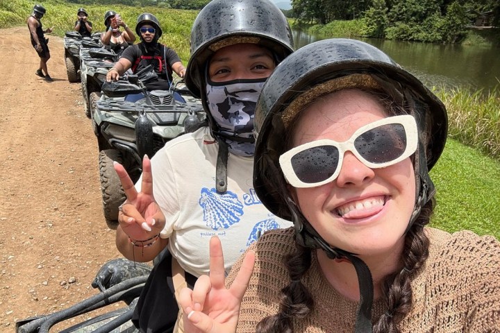 People wearing helmets on ATVs near a river, smiling and making peace signs.