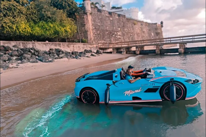 Blue car with 'Miami' logo driving on water near a stone fortress and lush trees.