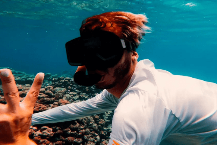 Person freediving underwater with diving mask, gesturing peace sign, colorful coral below.