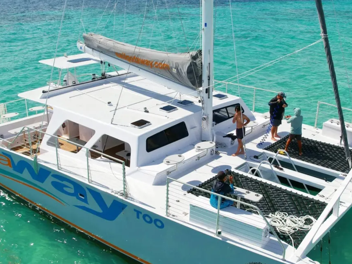People on a white catamaran in clear turquoise water, with a visible sail.