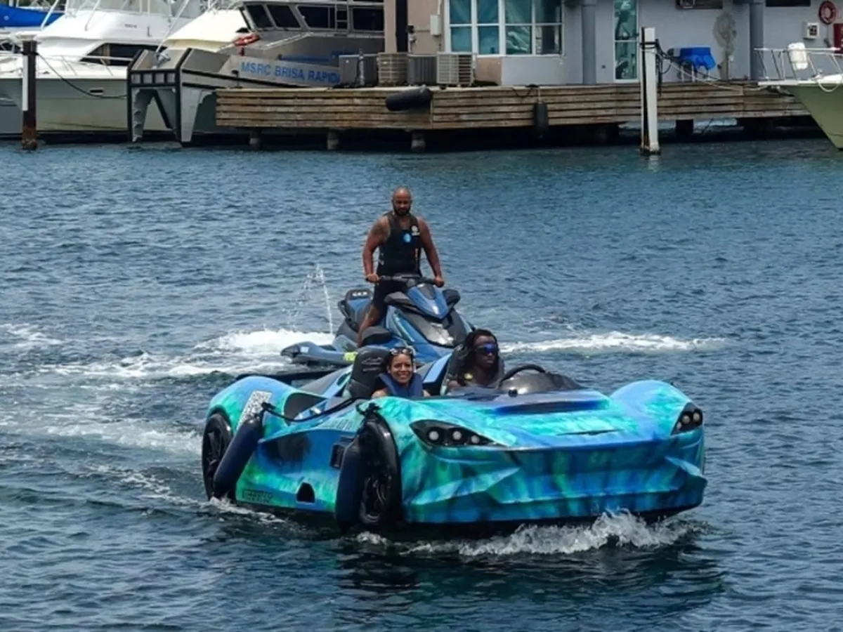 A car-shaped boat with people inside, followed by a man on a jet ski on the water.