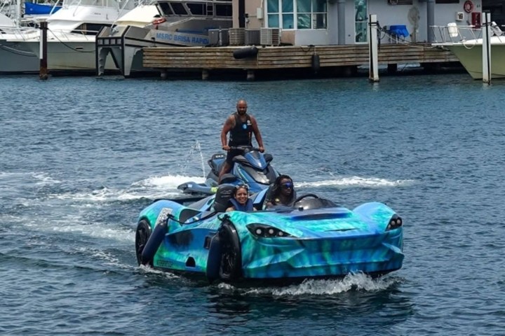 A car-shaped boat with people inside, followed by a man on a jet ski on the water.