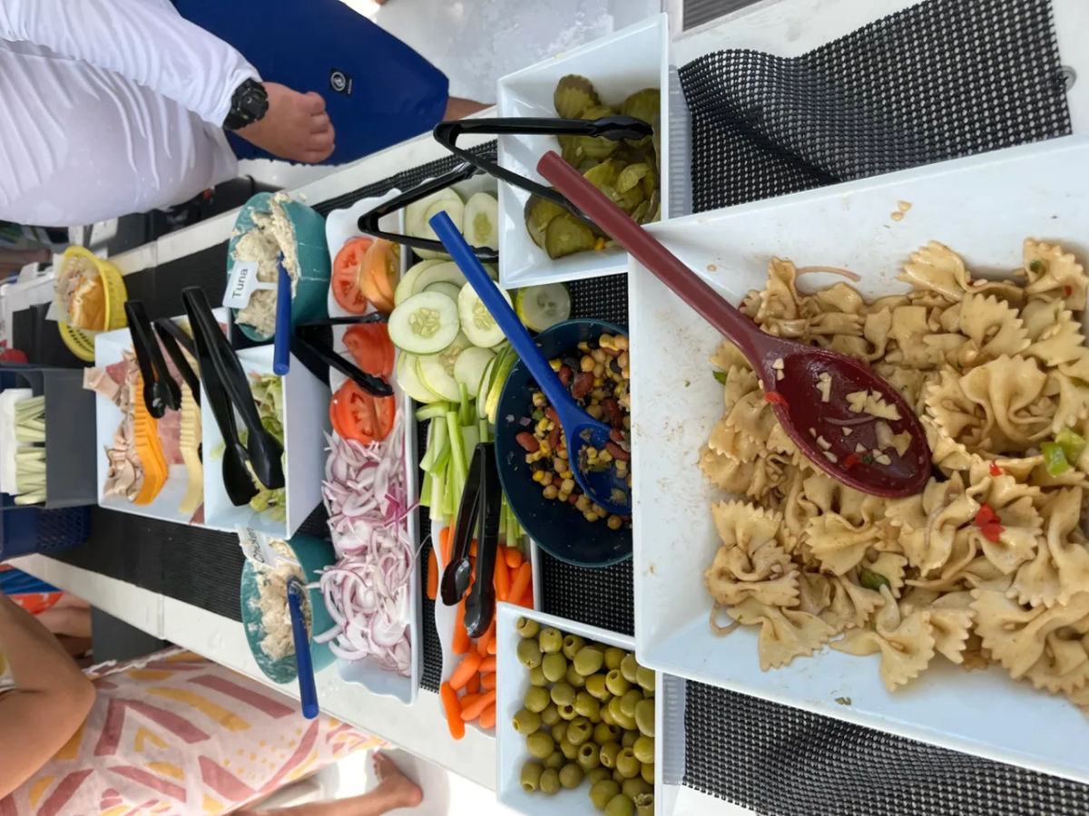 Buffet with pasta salad, vegetables, and deli meats on white trays.