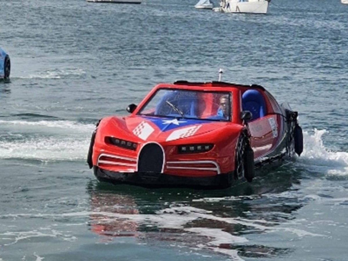 Amphibious car painted red and blue drives on water with boats in the background.