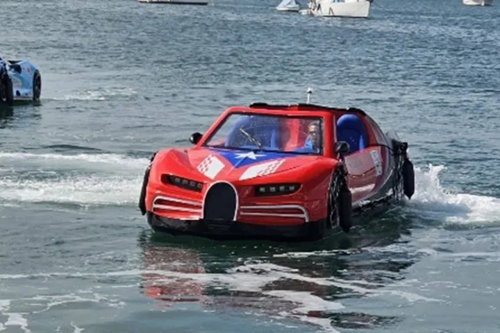 Amphibious car painted red and blue drives on water with boats in the background.