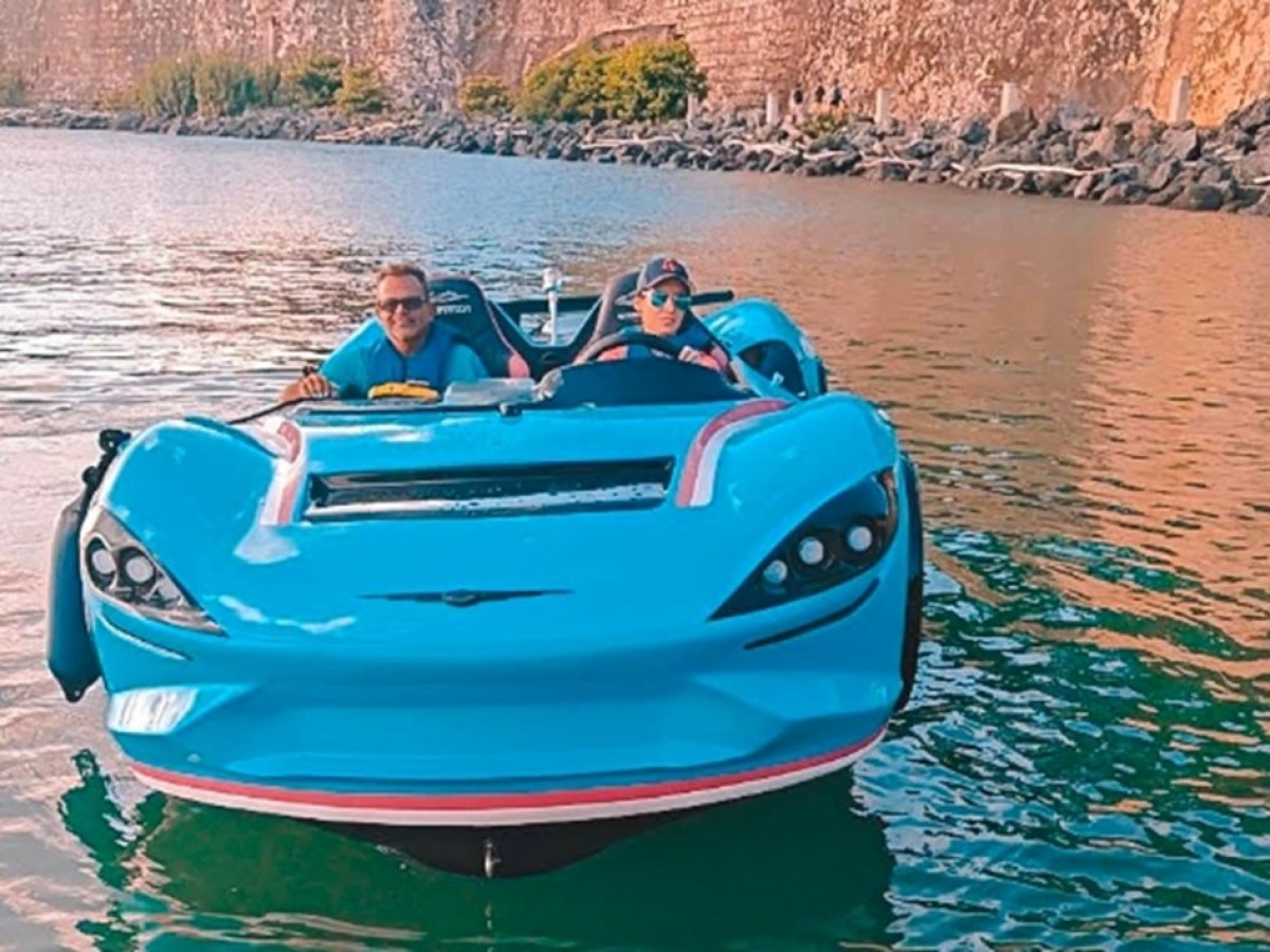 Two people in a blue floating car on a body of water near rocky cliffs.