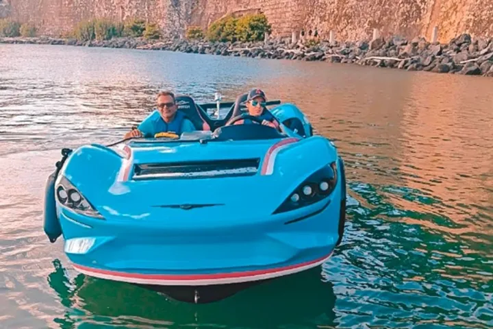 Two people in a blue floating car on a body of water near rocky cliffs.