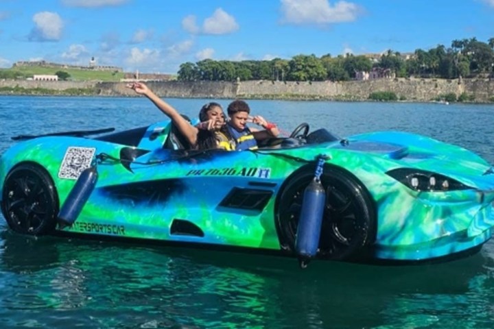 Two people in a colorful sports car shaped boat on water, with a scenic background.