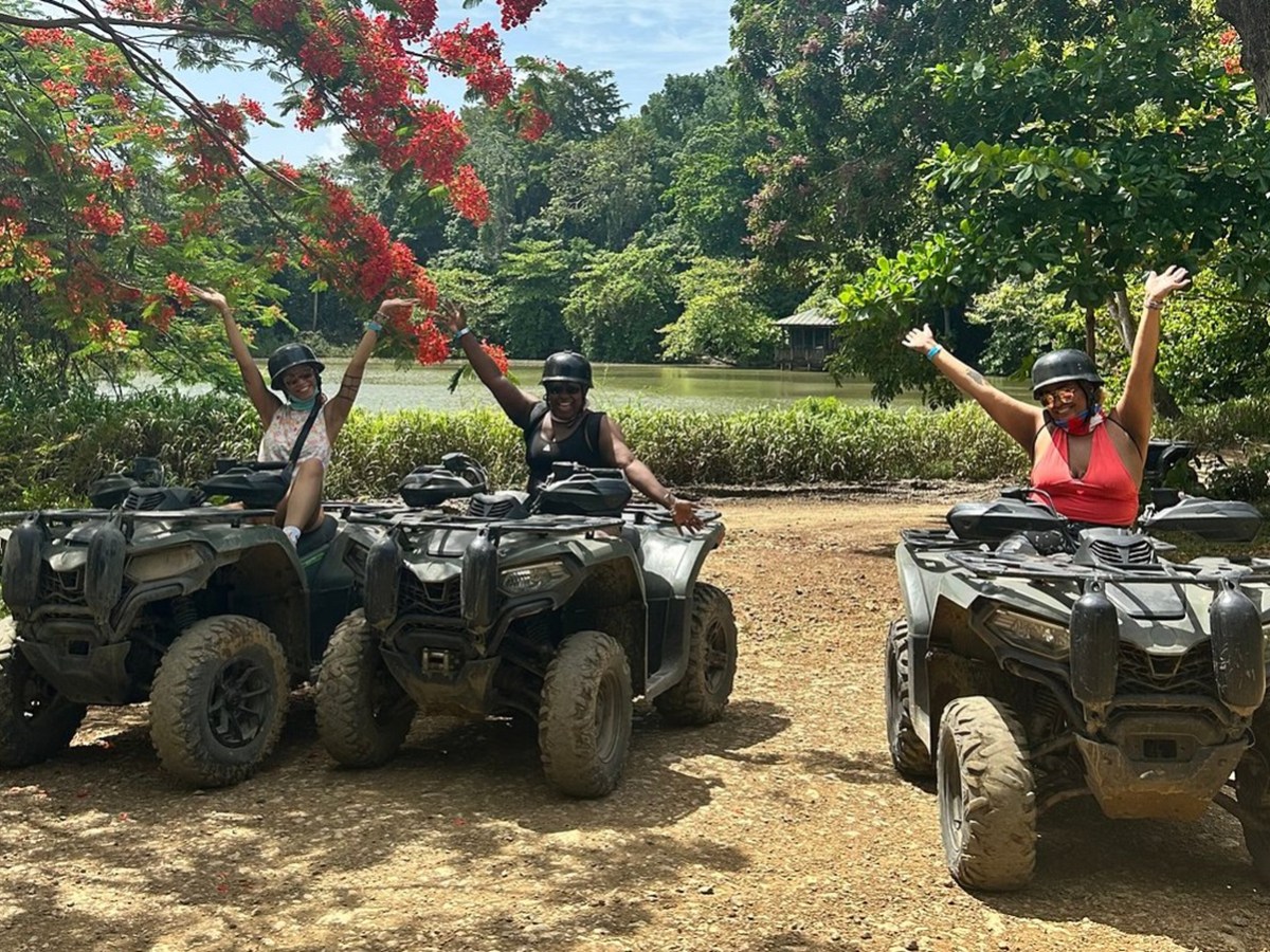 Three people on ATVs with arms raised, under trees with red flowers.