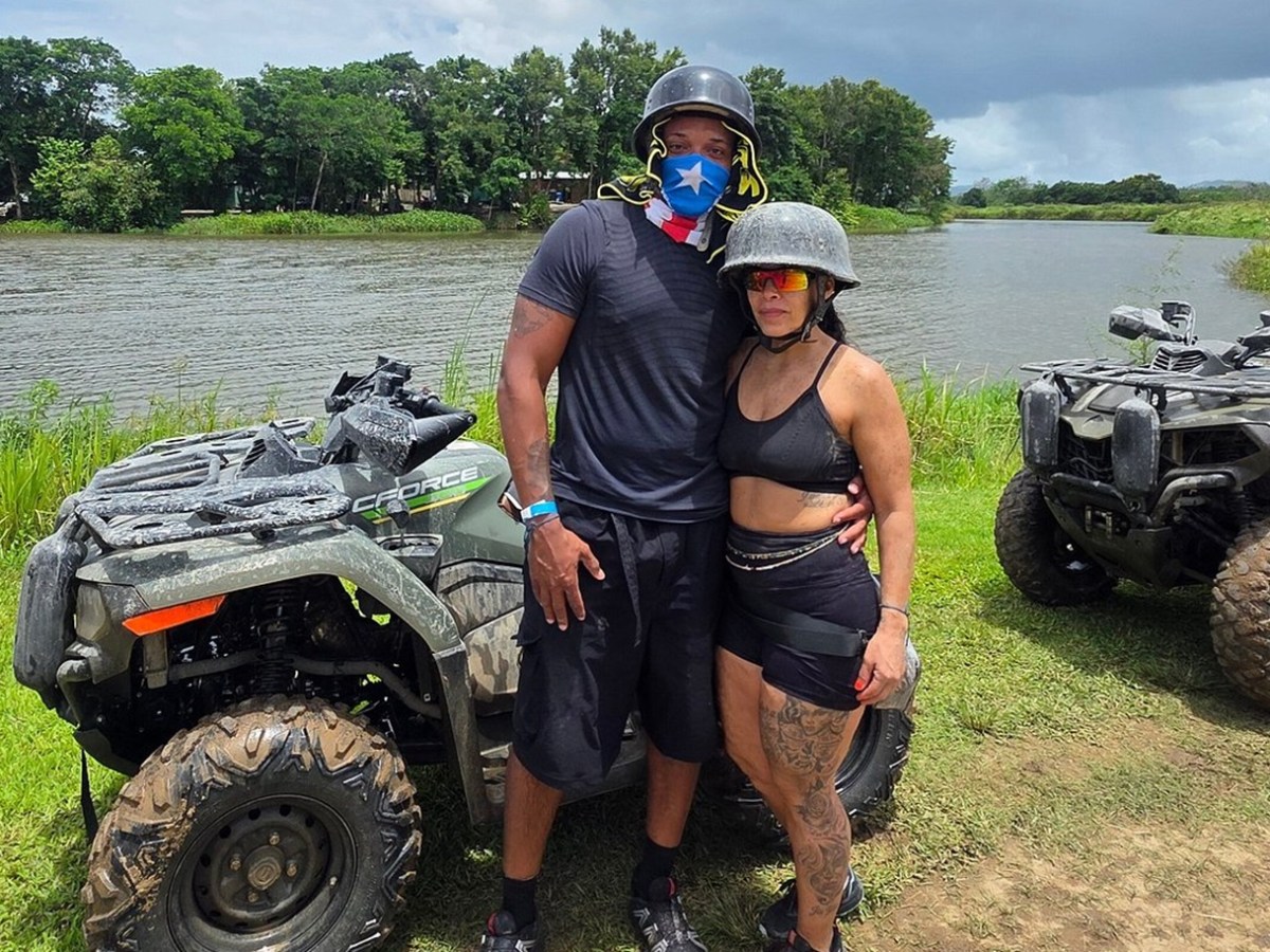 Two people in helmets by ATVs near a river with trees in the background.