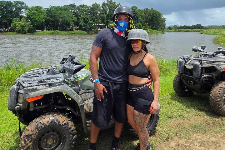 Two people in helmets by ATVs near a river with trees in the background.