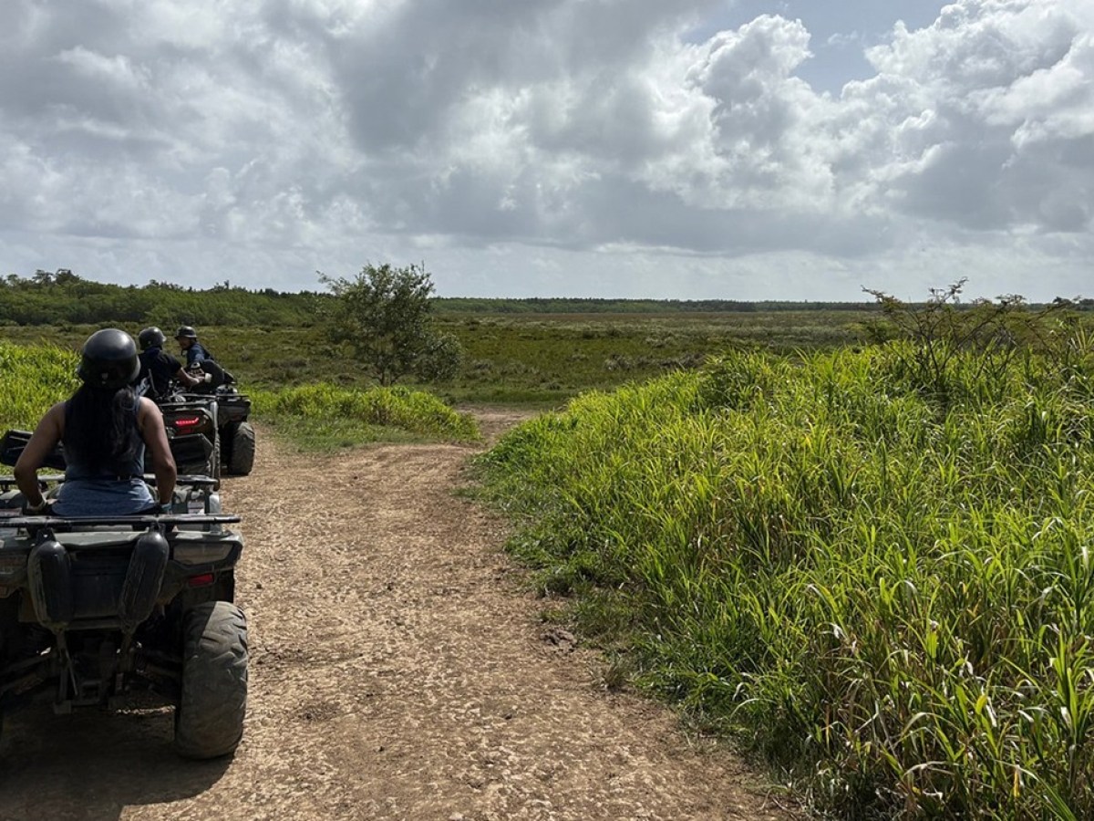 Two people riding ATVs on a dirt path through a grassy landscape under a cloudy sky.