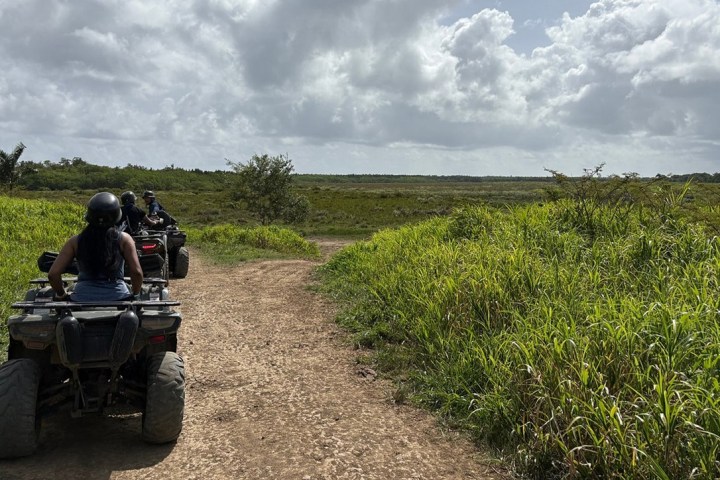 Two people riding ATVs on a dirt path through a grassy landscape under a cloudy sky.