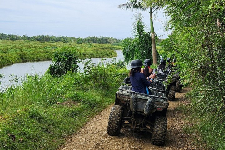 People riding ATVs on a dirt path beside a river in a lush, green landscape.