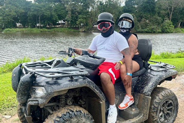 Two people wearing helmets sit on an ATV by a lake, surrounded by trees.