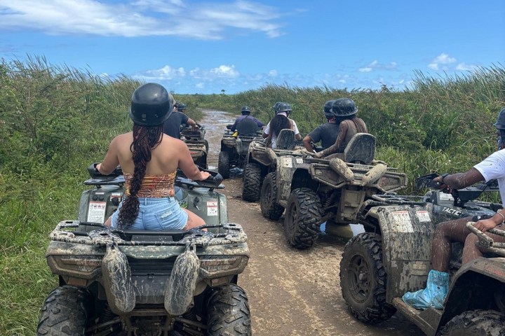 Group riding muddy ATVs on a dirt path surrounded by tall grass under a blue sky.