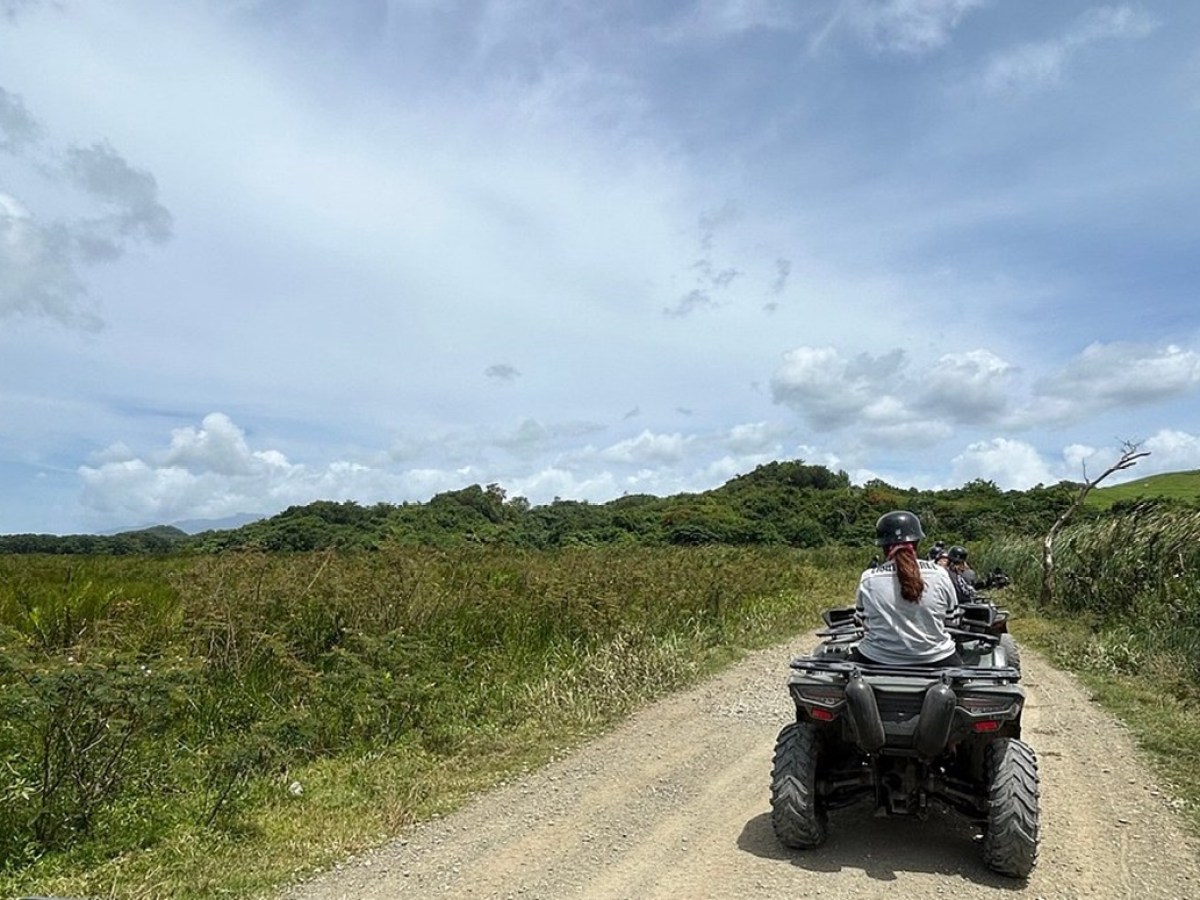 People riding ATVs on a dirt road through grassy landscape under a cloudy sky.