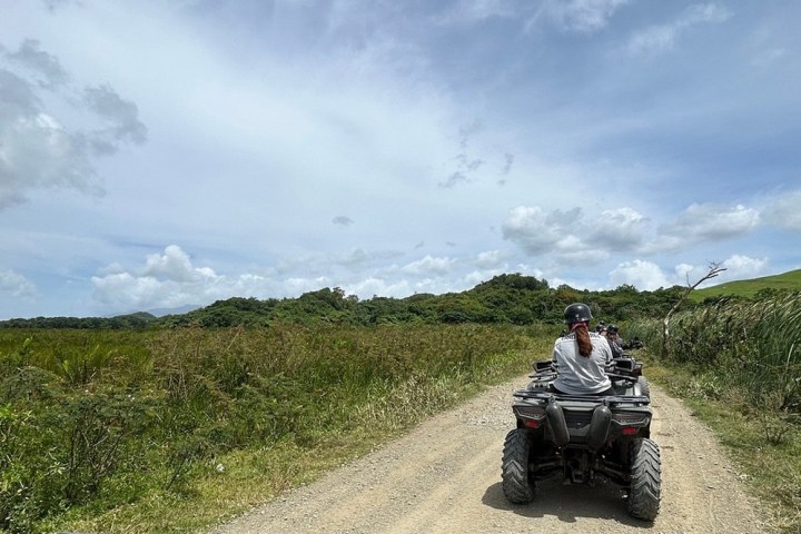 People riding ATVs on a dirt road through grassy landscape under a cloudy sky.