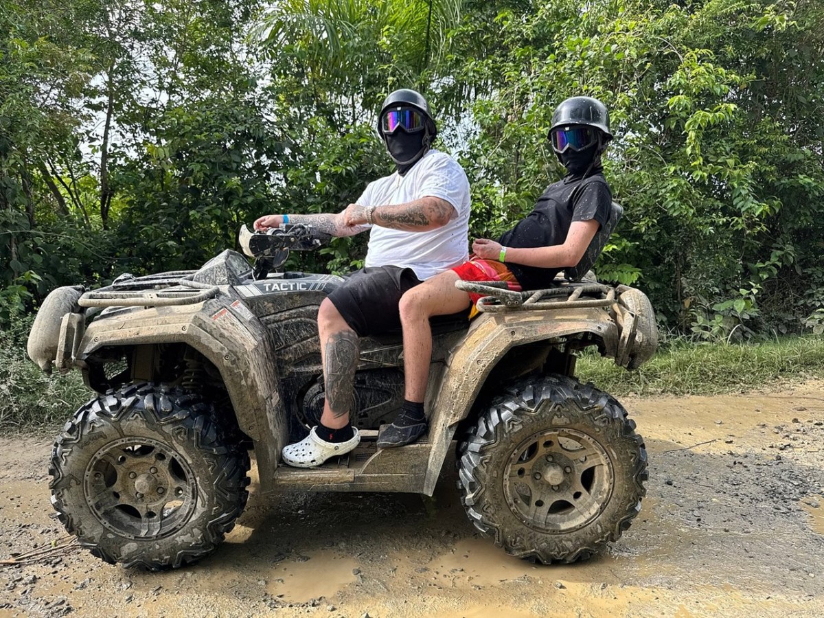 Two people ride a muddy ATV on a dirt path, wearing helmets and sunglasses.
